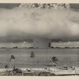May include: A black and white photograph of a mushroom cloud rising from the ocean. The cloud is large and billowing, and it is casting a shadow over the water. There are palm trees in the foreground, and a small island in the distance.