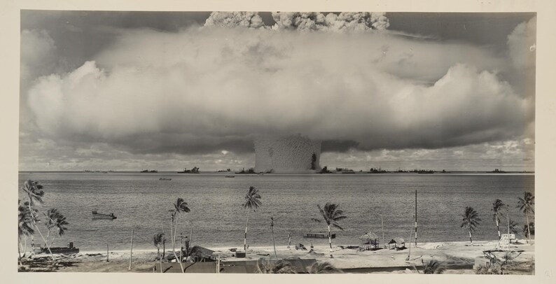 May include: A black and white photograph of a mushroom cloud rising from the ocean. The cloud is large and billowing, and it is casting a shadow over the water. There are palm trees in the foreground, and a small island in the distance.