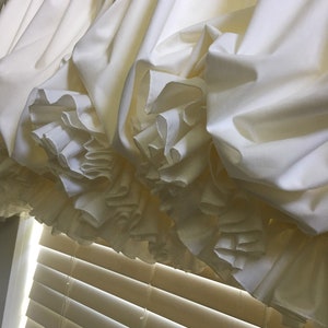 May include: A white ruffled valance with a gathered design, hanging above a set of wooden blinds.