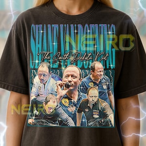 May include: Black t-shirt featuring a graphic of Shane Van Boening with text that reads "SHANE VAN BOENING" and "The South Dakota Kid." The shirt has a vintage, distressed look. The text at the bottom provides sizing guidance.