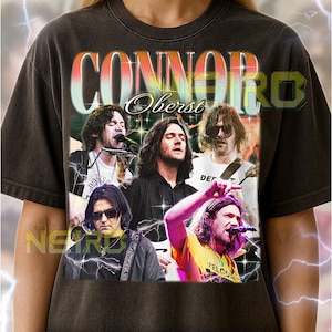 May include: Dark gray unisex t-shirt featuring a graphic of Connor Oberst and bandmates. The shirt has the band name in a retro font. The image includes the text "NEIRD" and "WELCOME".