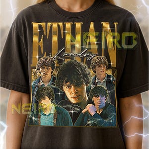 May include: Dark gray unisex t-shirt featuring a gold-colored graphic with the name "ETHAN Landry" and multiple images of a person. The shirt includes the text "NEW" and additional text at the bottom.