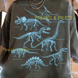 Peut inclure: T-shirt unisexe gris foncé avec des illustrations bleues de squelettes de dinosaures, dont Triceratops, Tyrannosaurus Rex et Brachiosaurus. Le t-shirt porte les inscriptions "TRICERATOPS", "ANKYLOSAURUS", "STEGOSAURUS", "PTERANODON", "BRACHIOSAURUS" et "ALLOSAURUS".