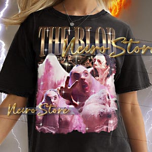 以下が含まれることがあります： 「THE BLOB Store」の文字が金色で描かれたグラフィックデザインの黒いTシャツ。デザインには、ピンクと白の複数の人物の画像が含まれています。モデルがTシャツを着ています。