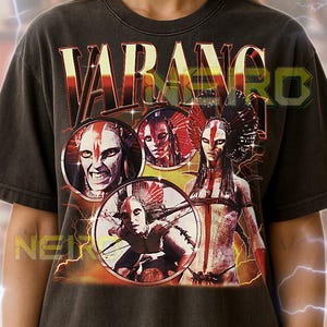 May include: Dark gray unisex t-shirt featuring the word "VARANGRO" in a bold, red and yellow font. The shirt displays circular portraits of figures with tribal face paint and headdresses. The text at the bottom provides sizing guidance.