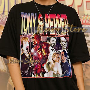 May include: Black unisex t-shirt featuring a graphic design with the text "TONY STARK POTUS" and images of characters from a movie. The design includes a red and gold Iron Man figure and other characters. The shirt is worn by a person.