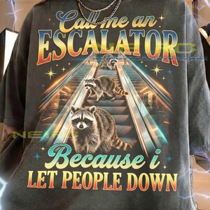 May include: Dark gray unisex t-shirt with the text "Call me an ESCALATOR Because i LET PEOPLE DOWN." The graphic features raccoons on an escalator with a retro design.