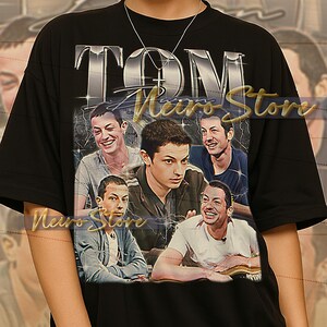 Pode incluir: Camiseta unissex preta com um gráfico de um homem e o texto prateado "TOM". A camiseta tem um design vintage com várias imagens do homem. O texto "Nero Store" também é visível.