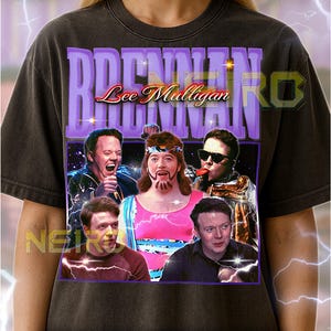 May include: Dark gray unisex t-shirt featuring a graphic of a comedy group with the text "BRENDAN Lee Mulligan" in purple. The image includes several people in various poses. The shirt has a vintage look.