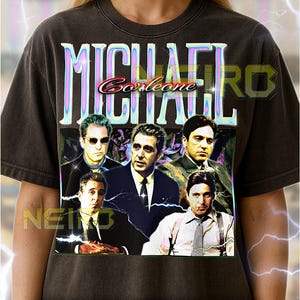 May include: Dark gray unisex t-shirt featuring a graphic of the movie "Michael Corleone" with multiple actors. The shirt has the text "MICHAEL Corleone" in a colorful, stylized font. The shirt is a vintage style.