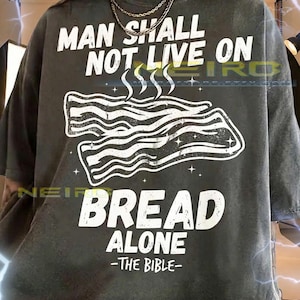 Op de afbeelding: Donkergrijs unisex T-shirt met de witte tekst "MAN SHALL NOT LIVE ON BREAD ALONE - THE BIBLE". De afbeelding toont een gestileerde afbeelding van spek. Een casual kledingstuk.