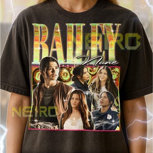 May include: Dark gray unisex t-shirt featuring a graphic design with the text "BAILEY" in large, colorful letters. The design includes images of women. The shirt has a vintage, distressed look.