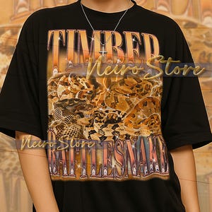 以下が含まれることがあります： ヴィンテージスタイルの「TIMBER RATTLESNAKE」の文字がプリントされた黒いTシャツ。茶色とベージュの色合いでガラガラヘビの詳細なイラストが描かれています。このシャツはユニセックススタイルです。