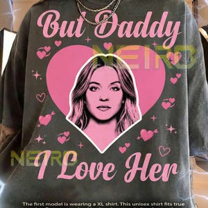 May include: Dark gray unisex t-shirt with the text "But Daddy I Love Her" in pink. The shirt features a pink heart with a woman's face in the center, surrounded by pink hearts and arrows.