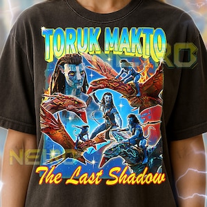 May include: Dark gray unisex t-shirt featuring a colorful graphic with the text "TORUK MAKTO" and "The Last Shadow." The design includes images of blue-skinned characters riding winged creatures, set against a blue and orange background.