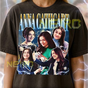 May include: Dark gray unisex t-shirt featuring a graphic of Anna Cathcart. The design includes multiple images of the actress with the text "ANNA CATHCART" in a blue and white font. The shirt is a vintage style.