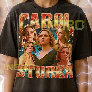 May include: Dark gray unisex t-shirt featuring a vintage-style graphic of a woman with the text "CAROL STURKA" in a retro font. The design includes multiple images of the woman. The shirt has a worn, distressed look.