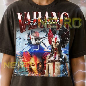 May include: Dark gray unisex t-shirt featuring the text "VADANG The Tsahik" in a stylized font. The graphic design includes three figures with elaborate headdresses and face paint, set against a blue and white background with lightning effects.