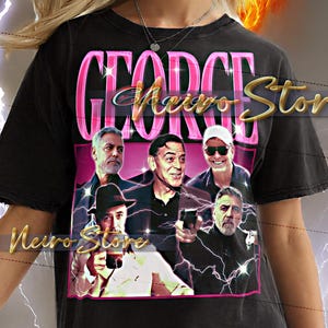 May include: Black unisex t-shirt with a graphic featuring the text "GEORGE" in pink and "Meiro Stor" in gold. The graphic includes images of actors. The shirt is worn by a person.