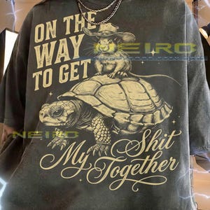 Puede incluir: Una camiseta gris carbón con el gráfico de un ratón con sombrero de vaquero montado sobre una tortuga. El texto dice ON THE WAY TO GET My Shit Together. El diseño está impreso en color crema claro con pequeños detalles de estrellas.