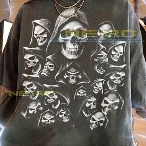 May include: A charcoal gray short-sleeve t-shirt featuring a large graphic print of multiple hooded skulls. The text NEIRO is printed in yellow across the center. The bottom includes text regarding sizing and fit for this unisex garment.