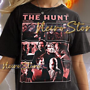May include: Black unisex t-shirt featuring a movie poster design for "The Hunt" with Mads Mikkelsen. The shirt includes the film's title, cast, and year. The model is wearing a 2XL shirt.