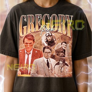 Puede incluir: Camiseta unisex gris oscuro con un diseño gráfico vintage de Gregory Peck. El diseño incluye el nombre "GREGORY Peck" en una fuente retro, junto con múltiples imágenes del actor. La camiseta tiene un cuello redondo clásico.