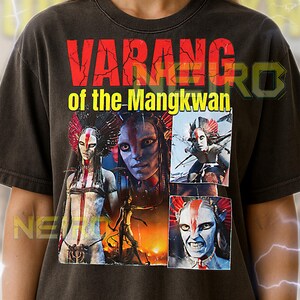 May include: Dark gray unisex t-shirt featuring the text "VARANG of the Mangkwan" in red and yellow. The shirt displays multiple images of a character with blue skin and tribal markings. The shirt is made of a soft material.
