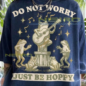 May include: Navy blue t-shirt with a whimsical design featuring frogs. The shirt has the text "DO NOT WORRY" above a frog playing a guitar, and "JUST BE HOPPY" below. Musical notes and stars accent the design.