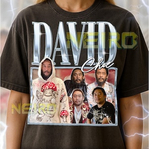May include: Dark brown unisex t-shirt featuring the name "DAVID" in a metallic silver font, with "Choe" in script. The shirt displays a framed image of several men. The text "NEA" is visible on the left side of the shirt.