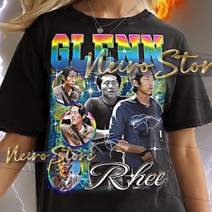 May include: Black t-shirt featuring a graphic of Glenn Rhee from The Walking Dead. The design includes multiple images of the character and the name "Glenn Rhee" in a colorful, retro style. The shirt is worn by a person.