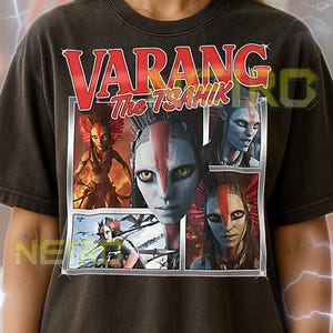 May include: Dark gray unisex t-shirt featuring the text "VARANG The TSAHIK" in red and white. The shirt displays a multi-panel graphic of a character with blue skin, red markings, and a feathered headdress. The shirt is a unisex fit.