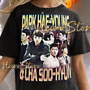 May include: Black t-shirt featuring a graphic of several people with the text "PARK HAE-YOUNG" and "CHA SOO-HYUN". The shirt has a distressed look and is worn by a person with blonde hair.
