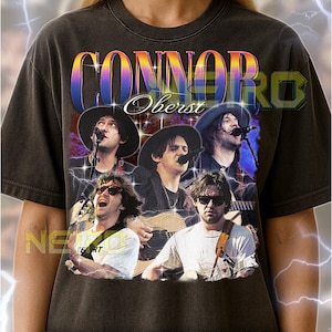 May include: Dark gray unisex t-shirt featuring a band photo with the text "CONNOR OBERST" in a colorful, retro font. The shirt has a vintage look and feel, with a distressed graphic design.
