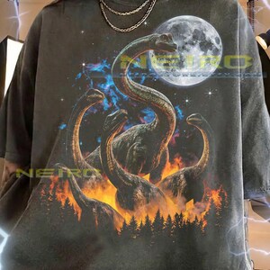 Peut inclure: T-shirt unisexe gris foncé avec un motif de dinosaures au long cou, sur fond de pleine lune, de flammes et de ciel étoilé. Le t-shirt comprend le texte : "The first model is wearing a XL shirt. This unisex shirt fits true to size."