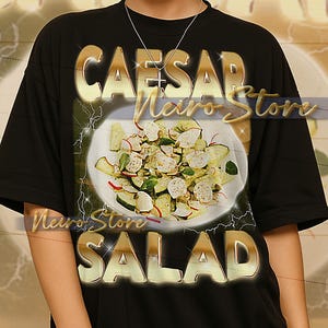 Puede incluir: Camiseta unisex negra con letras doradas "CAESAR SALAD". La camiseta presenta un gráfico de ensalada. La modelo lleva una talla XL. También se ve el texto "Neiro Store".