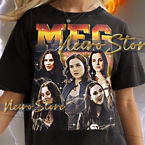 Pode incluir: T-shirt preta com um design gráfico dourado com a palavra "MEG" e imagens de mulheres. A camisa tem um visual vintage e desgastado. O texto "Neiro Store" também é visível.