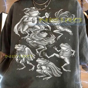 Op de afbeelding: Donkergrijs unisex T-shirt met een gedetailleerde illustratie van verschillende antropomorfe kikkers in capes en vesten. De kikkers staan in verschillende poses, sommige houden muziekinstrumenten vast. De tekst onderaan geeft maat informatie.