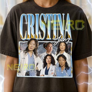 May include: Dark gray unisex t-shirt featuring a graphic of the character Cristina Yang from the TV show. The design includes the name "Cristina Yang" in a stylized font and images of the character. The shirt also has text at the bottom.