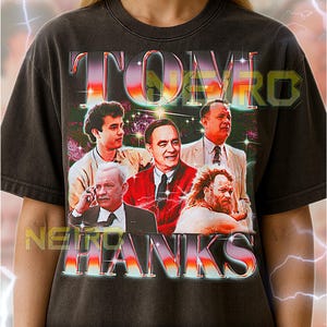 May include: Dark gray unisex t-shirt featuring a retro design with the text "TOM HANKS" in a colorful, 3D font. The graphic includes a collage of images of Tom Hanks and other actors. The shirt fits true to size.