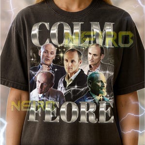 May include: Dark gray unisex t-shirt featuring a graphic with the text "COLMO NADORE" in silver metallic letters. The graphic includes images of several men and lightning bolts.