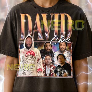 May include: Dark gray unisex t-shirt featuring a graphic of the name "DAVID" in gold lettering above a framed image of several men. The image includes the name "Choe" in script. The shirt has text at the bottom.