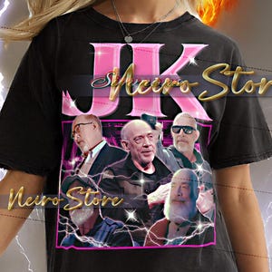 May include: Black t-shirt featuring a graphic design with the text "JK Neuro Store" in pink and gold. The design includes images of several men. The model is wearing a 2XL shirt.
