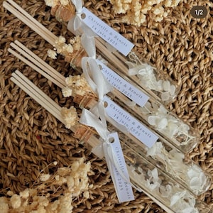 Può includere: Quattro piccole bottiglie di vetro con tappi di sughero riempite di fiori bianchi secchi e legate con un nastro bianco. Ogni bottiglia ha un'etichetta bianca con il testo "Christelle & Benoit 14 giugno 2025".