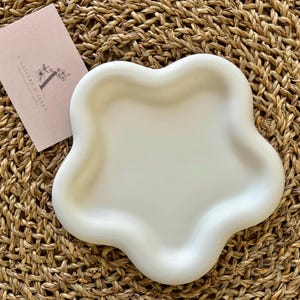 Può includere: Un vassoio in ceramica bianca con un design unico a forma di fiore organico. Il vassoio è su una superficie intrecciata.