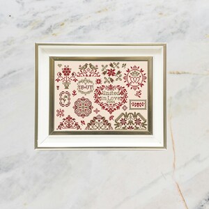 Pode incluir: Amostra de ponto cruz emoldurada com fundo creme e uma variedade de desenhos florais e geométricos bordô e verde-oliva. O texto "United in Love 2011" está presente. A moldura é prateada.
