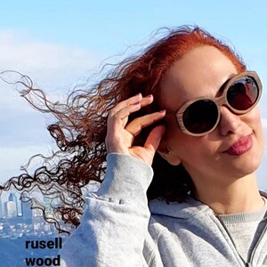 Könnte beinhalten: Eine Frau mit langen, lockigen roten Haaren trägt eine braune Sonnenbrille mit Holzrahmen und dunklen Gläsern. Sie trägt ein graues Kapuzenpullover und ist im Freien mit einer Skyline der Stadt im Hintergrund.