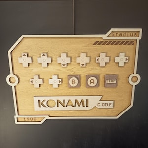 Könnte beinhalten: Eine Wandtafel aus hellem Holz mit der Konami-Code-Sequenz, bestehend aus Richtungskreuz-Symbolen sowie B-, A- und Start-Tasten. Der Text Gradius, Konami Code und 1986 ist in die Oberfläche eingraviert. Das Design hat einen geometrischen Rand.