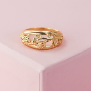 May include: A gold ring with a delicate, intricate design featuring a floral motif. The ring is made of a smooth, polished metal and has a wide band.