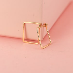 Könnte beinhalten: Ein Paar goldfarbene quadratische Creolen. Die Ohrringe werden auf einer rosa Oberfläche neben einer rosa Schachtel präsentiert. Die Ohrringe haben ein schlichtes, geometrisches Design und ein glänzendes Finish.